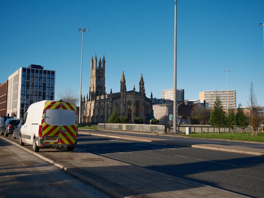 A white delivery van with yellow and red reflective markings on the rear windows is parked on the street near a pavement, close to the entrance of a historic church with tall, ornate spires. The van is positioned next to a row of parked cars, and the scene is captured in bright daylight with a clear blue sky. Modern buildings surround the church, and the area appears to be part of an urban environment. The street is lined with street lamps and small trees, with some greenery visible in front of the church. This image illustrates a typical scene in a city area, potentially involving home relocation or furniture transport, with the van likely used for removal or moving services. [COMPANY_NAME] may use such vehicles for assisting with packing, loading, and transporting household items during a house removal process on Grange Road, Riddlesdown, as highlighted in the page titled 'Avoid Van Access Problems on Grange Road, Riddlesdown.'