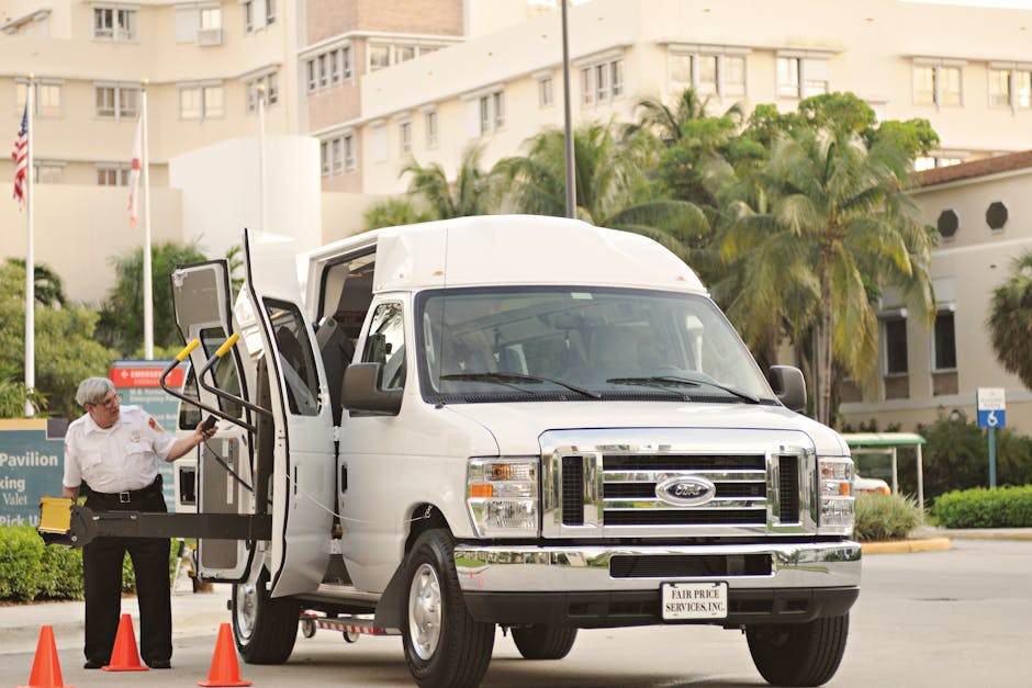 A white commercial van parked on the street in front of a red brick building with decorative window frames. The van is positioned parallel to the curb, with the driver's side facing the sidewalk. The vehicle appears to be part of a home relocation or furniture transport process, commonly used by removals services such as Man with Van Riddlesdown. The street surface is asphalt, and there are a few pedestrians walking along the sidewalk. Bright natural lighting enhances the visibility of the van's details, including its side panels, wheels, and the black trim along the lower portion. The surrounding environment suggests an urban setting suitable for loading or unloading household items during a house removal or move, with the van ready for a loading process or just completing transportation of packed boxes and furniture.