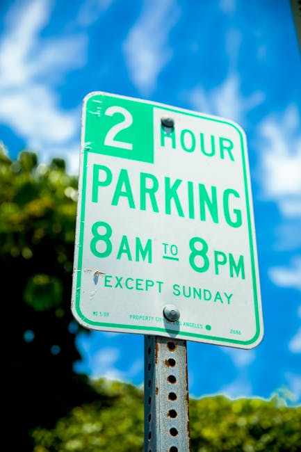 Close-up of a green and white municipal parking sign mounted on a metal pole in an outdoor setting. The sign indicates a two-hour parking limit from 8 am to 8 pm, Monday through Saturday, with an exception for Sundays. The background features a bright blue sky with scattered white clouds and areas of greenery, including trees with lush foliage. The sign is positioned in a residential or urban area, likely near a property involved in house removals or moving services, as suggested by the context from manwithvanriddlesdown.co.uk. The image captures the sign during daytime with natural lighting, emphasizing clear visibility and readability of the parking regulations, relevant for planning logistical aspects of home relocation or furniture transport in Riddlesdown, Croydon.