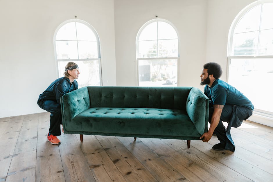 Two men are inside a bright, spacious room with white walls and large arched windows allowing natural light. They are lifting a green velvet sofa, holding it firmly at each end to prepare for moving or furniture transport as part of a home relocation or packing and moving process. One man has light skin and long hair tied back, dressed in a dark blue top and red sneakers, while the other has dark skin, an afro hairstyle, and wears a matching dark blue top with black trousers. The sofa is positioned across a wooden floor, and behind the men, there are no other visible objects or furniture. The room appears clean and minimally furnished, with the focus on the lifting action, indicative of furniture loading or unloading for a removal service. This visual captures the careful manual handling involved in furniture removals, supported by the professional services of Man with Van Riddlesdown, ensuring efficient and safe furniture transport during house removals or moving logistics.