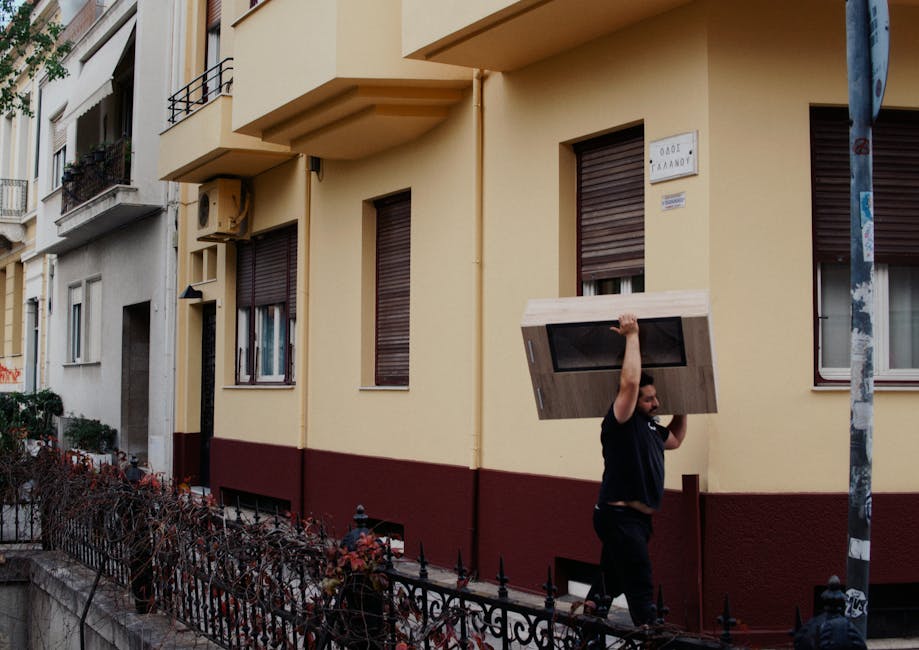 A man wearing a black shirt is lifting a large piece of flat-packed furniture, likely a wooden cabinet or wardrobe, over his shoulder as he walks on a pavement outside a yellow residential building with dark brown window shutters. The furniture is wrapped in protective packaging, such as plastic or fabric, to prevent damage during transport. The building features multiple windows with closed shutters, and a small sign is affixed to the wall, indicating an address or nameplate. The man appears to be engaged in a home relocation process facilitated by Man with Van Riddlesdown, part of a moving service specializing in furniture transport and packing. The scene shows the activity happening during daylight, with a chain-link fence and some plants bordering the sidewalk in the foreground, and a street sign partially visible on the right. This image captures the logistical aspect of loading furniture onto a vehicle in preparation for a house move, with emphasis on careful handling and the transport process.