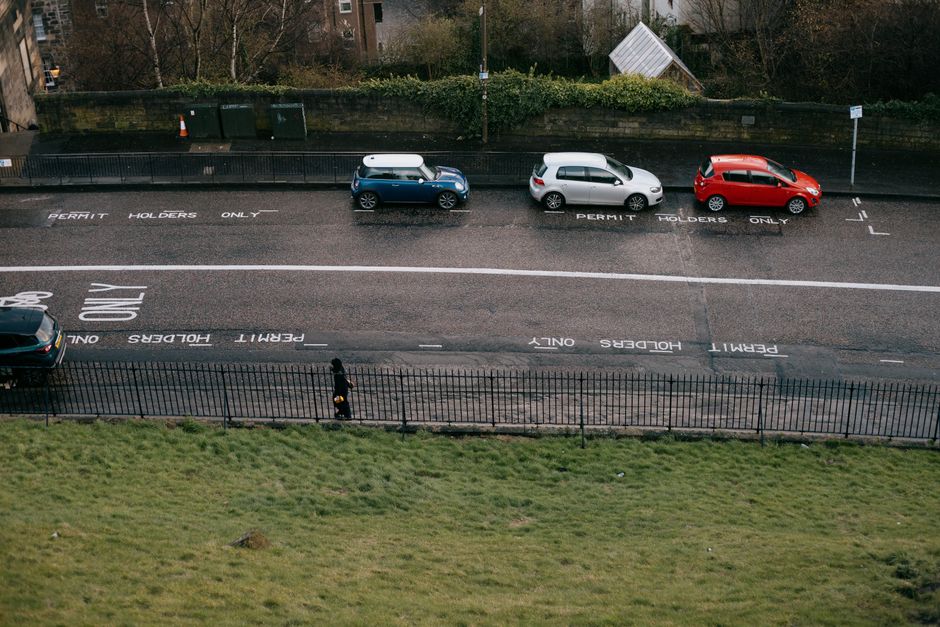 An aerial view of a parking area on Riddlesdown Lane showing three cars parked parallel along the curb, including a blue compact vehicle, a silver hatchback, and a red hatchback. The parking bays are marked with white lines and labeled with instructions for permit holders only, with the words 'PERMIT HOLDERS ONLY' painted on the asphalt. Behind the cars, there is a low brick wall topped with green hedges, and a few traffic cones are positioned near the wall. In the foreground, a black wrought iron fence runs horizontally, separating the parking area from a grassy field below. A person dressed in dark clothing is walking along the pavement, adjacent to the fence. The parking lot appears clean and organized, with no visible moving equipment or furniture, indicating a typical street-side parking scene in a residential or urban area where house removals and furniture transport could take place. Man with Van Riddlesdown occasionally operates in such environments for local relocations and packing logistics.