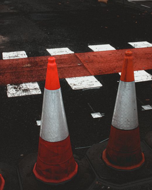 Two orange and white traffic cones placed on a paved street surface, with a red painted section and white dashed lines indicating a pedestrian crossing or road marking behind them. The cones are positioned near the curb, adjacent to a designated loading or loading zone area, which is part of the home relocation process. The scene captures an outdoor environment relevant to vehicle access and street management, as seen in urban or suburban areas where removals and furniture transport are carried out by [COMPANY_NAME]. The lighting suggests daytime, and the cones serve to manage traffic or restrict vehicle movement in the vicinity of a property, supporting the logistical aspects of house removals and moving services.