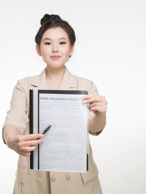 A professional woman with neatly styled dark hair, wearing a beige blazer, standing against a plain white background while holding a clear plastic folder with a visible 'Home Insurance Policy' document inside. She is presenting the document using both hands, one at the top and the other at the bottom, with a black pen in her left hand pointing towards the paper. The image conveys a sense of preparedness and assurance, relevant to house removals and home relocation services provided by Man with Van Riddlesdown, highlighting careful planning and insurance coverage for moving projects. The lighting is even and neutral, focusing on the woman and the document, emphasizing the importance of insurance during furniture transport and packing during the house moving process.