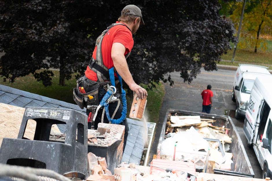 A man wearing a grey cap, red T-shirt, and a safety harness is standing on the pitched roof of a residential building during a home relocation process. He is holding a brick in his right hand, preparing to load it into a rubbish skip that contains various debris, including wooden planks, cardboard, and plastic wrapping. The scene is illuminated by natural daylight, with large trees and a street visible in the background. Nearby, parked white vans are positioned for loading furniture and packing materials. The man with van Riddlesdown occasionally uses equipment such as straps and blankets during furniture transport and packing and moving tasks, which are typical for comprehensive removals services provided by [COMPANY_NAME]. The image captures the careful handling of building materials and the logistical aspects of the loading process, emphasizing the planning involved in professional house removals and relocation projects.