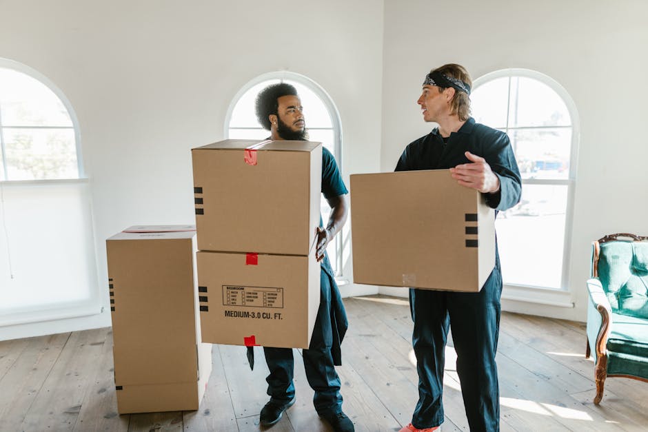 Two men involved in house removals inside a well-lit room with wooden flooring and white walls, preparing for a home relocation. One man, with a beard and curly hair, is holding a medium-sized cardboard box, while the other, wearing a headband, is carrying an empty box and gesturing as if discussing the packing process. Several other cardboard boxes, some labeled with size and volume details, are stacked nearby on the wooden floor. The room features two large arched windows allowing natural light to illuminate the space. A vintage-style green armchair with wooden accents is positioned against the wall on the right, providing context for a domestic interior. The scene captures the furniture transport and packing activities typical of a professional removal service, with the men wearing casual clothing suitable for moving tasks, supported by [COMPANY_NAME], highlighting the detailed loading and packing preparations involved in local moves from Riddlesdown to Purley.