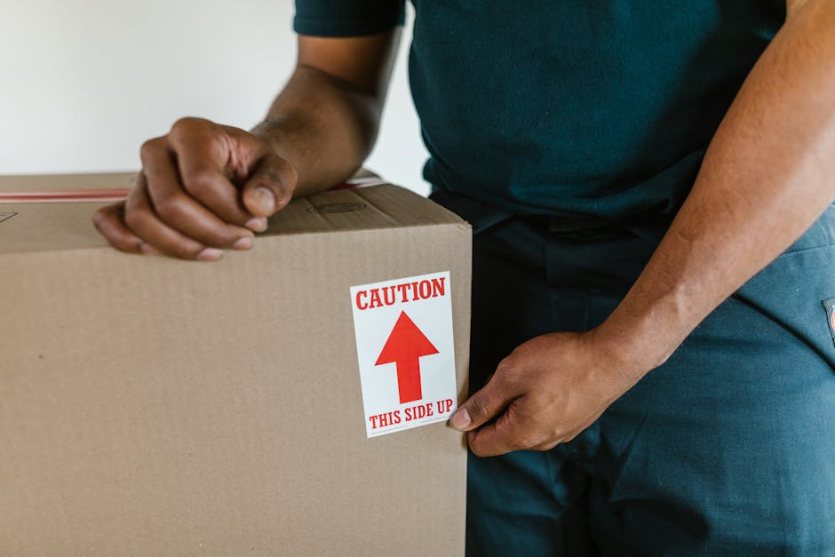 A person wearing a dark green T-shirt and dark blue work trousers is holding a medium-sized cardboard moving box with both hands, gripping the sides of the box. The box is positioned inside a home, likely near an entrance or a corridor, with a neutral-colored wall visible in the background. The box has a white label with red text and an upward arrow that reads 'CAUTION THIS SIDE UP,' indicating proper handling during packing and transport. The scene depicts the process of home relocation or furniture transport as part of a professional removals service by [COMPANY_NAME], with the individual preparing the box for loading into a van or for moving within the property. The environment is well-lit, emphasizing the importance of careful handling and loading procedures involved in professional house removals.