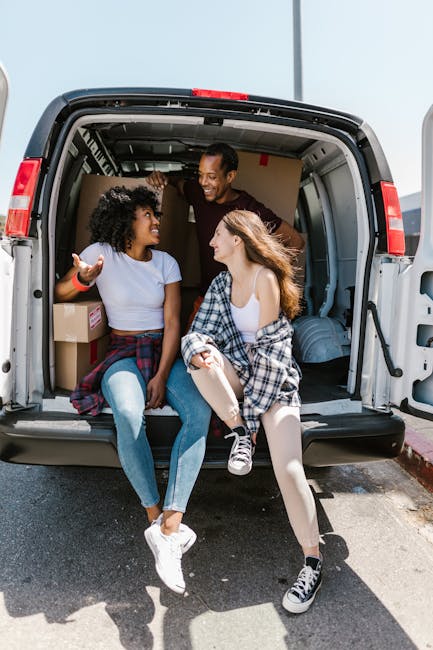 Two young women sitting inside the open rear of a moving van, parked on a driveway, during a home relocation. The woman on the left has curly dark hair, wearing a light blue t-shirt and jeans, with a cardboard box behind her. The woman on the right has long, wavy brown hair, wearing a white top, a plaid shirt tied around her waist, and light-colored leggings. Both are smiling and engaging in conversation, with the woman on the right holding her hair. The van's interior shows packed cardboard boxes and protective padding, indicating furniture transport and packing materials used during a move. The scene is illuminated by natural daylight, and the driveway appears clean and organized, reflecting typical moving logistics and loading processes associated with professional removals by Man with Van Riddlesdown.