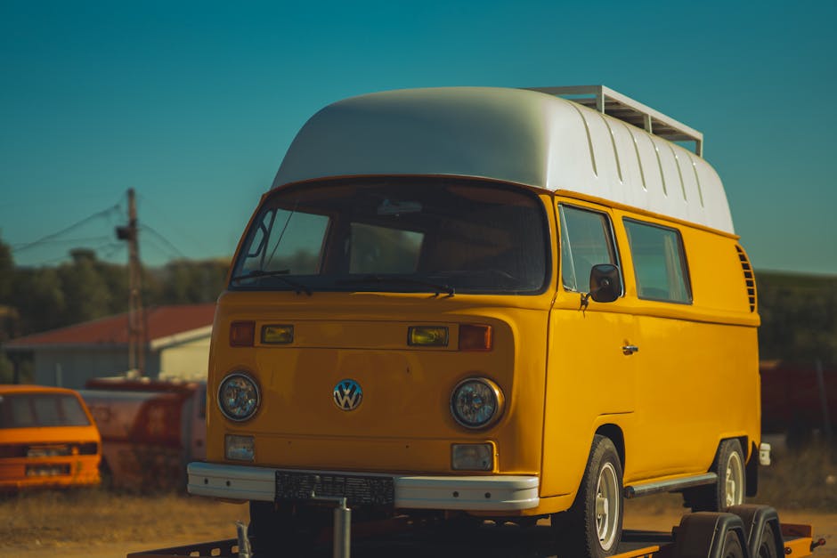 A vintage yellow Volkswagen Type 2 van with a white rounded roof parked outdoors on a mobile platform, with visible loading straps securing it. The van's front features a large windshield, round headlights, and the VW emblem in the center. In the background, there are other vehicles, including an orange van, and some structures and electrical poles under a clear blue sky. The scene is illuminated by natural daylight, suggesting daytime hours. This image relates to home relocation services provided by Man with Van Riddlesdown, illustrating furniture transport and vehicle loading processes involved in house removals, with the van possibly being prepared for transport or as part of packing and moving activities.