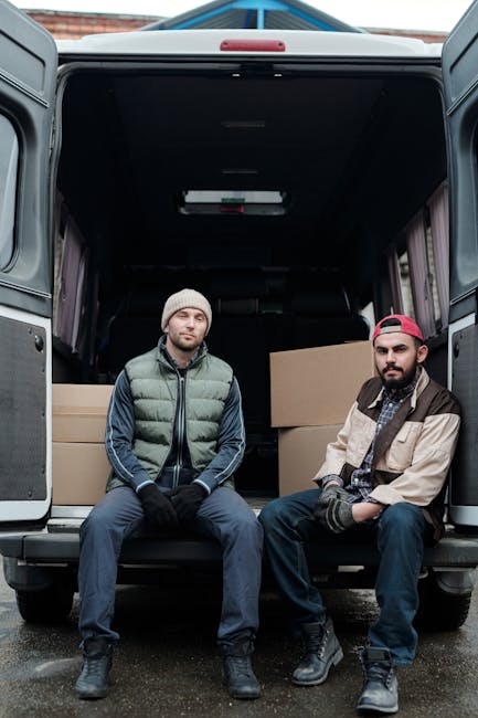Two young women sitting inside the open rear of a moving van, parked on a driveway, during a home relocation. The woman on the left has curly dark hair, wearing a light blue t-shirt and jeans, with a cardboard box behind her. The woman on the right has long, wavy brown hair, wearing a white top, a plaid shirt tied around her waist, and light-colored leggings. Both are smiling and engaging in conversation, with the woman on the right holding her hair. The van's interior shows packed cardboard boxes and protective padding, indicating furniture transport and packing materials used during a move. The scene is illuminated by natural daylight, and the driveway appears clean and organized, reflecting typical moving logistics and loading processes associated with professional removals by Man with Van Riddlesdown.
