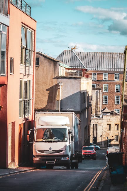 A white box truck with a closed cargo area is parked at the top of a narrow urban street adjacent to residential buildings, with some windows and brickwork visible. The truck is positioned on a slight