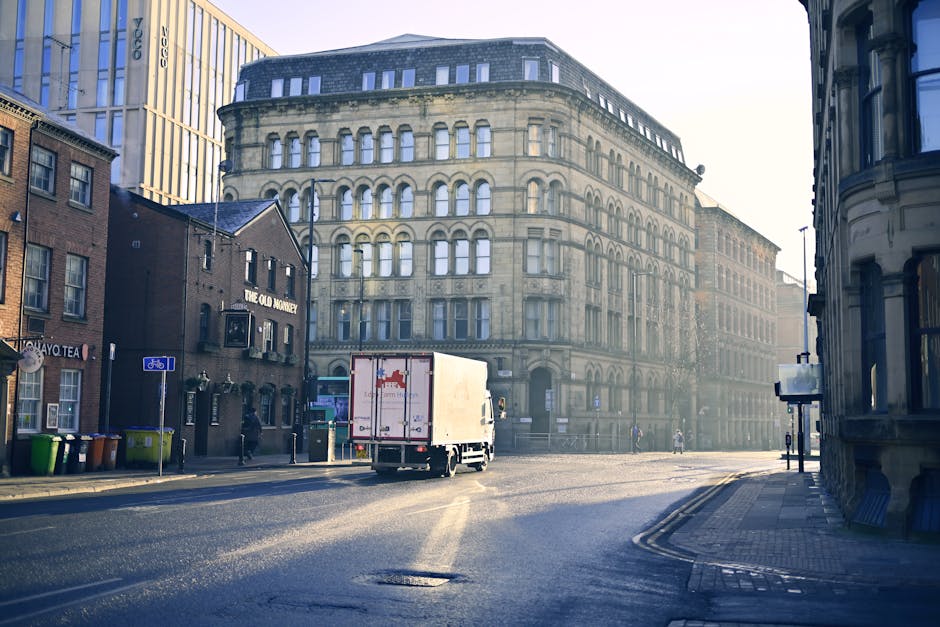 A white box truck with a closed cargo area is parked at the top of a narrow urban street adjacent to residential buildings, with some windows and brickwork visible. The truck is positioned on a slight