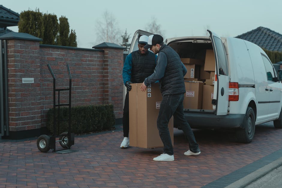 Two men engaged in a home relocation task outdoors, loading a large cardboard box into the back of a white moving van parked on a brick driveway. One man, wearing a black cap, black quilted vest, and grey pants, is lifting the box, while the other, dressed in a dark jacket and black gloves, helps guide it into the vehicle. The open van reveals neatly stacked cardboard boxes inside, some sealed with packing tape. Nearby, a black hand truck or dolly is positioned on the driveway, ready for use. In the background, a brick wall with a small hedge and residential houses with tiled roofs are visible under overcast skies. The scene illustrates the process of furniture transport and packing during a house move, with safety and efficiency in mind, typical of the services provided by Man with Van Riddlesdown, supporting local relocation and removals.