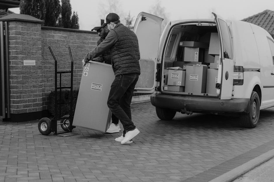 Two movers from Man with Van Riddlesdown are engaged in the loading process for a home relocation, transferring a large cardboard box with fragile contents onto a hand truck. They are outside a residential property with a paved driveway, next to a brick wall and a small garden. The open rear doors of a white delivery van reveal several packed cardboard boxes of various sizes inside, some labeled and wrapped in plastic packaging for transport. One mover, wearing a baseball cap and casual clothing, is lifting the box, while the other, dressed in a jacket and jeans, supports the load. The scene is set in natural daylight, highlighting the coordinated effort involved in packing and moving household items, with equipment such as a trolley, straps, and blankets occasionally used during the furniture transport and packing process by Man with Van Riddlesdown.