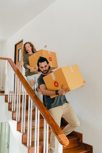 A man and a woman are carrying cardboard moving boxes down a staircase inside a residential property. The man, dressed in a dark t-shirt and beige trousers, is leading with two boxes, while the woman, wearing a green top and ripped jeans, follows behind holding another box. The staircase has wooden steps and a wooden handrail, with white walls surrounding it. The scene captures the loading process during a home relocation, supported by a professional removal service such as Man with Van Riddlesdown, involving careful packing, furniture transport, and staircase navigation inside the apartment.