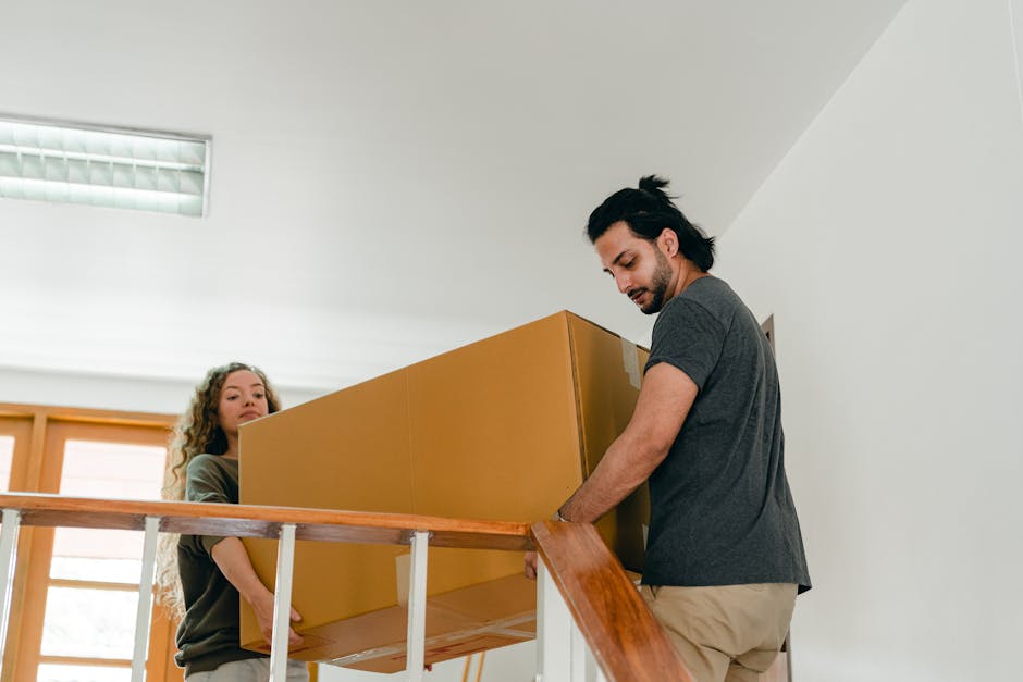 A man and a woman are carrying cardboard moving boxes down a staircase inside a residential property. The man, dressed in a dark t-shirt and beige trousers, is leading with two boxes, while the woman, wearing a green top and ripped jeans, follows behind holding another box. The staircase has wooden steps and a wooden handrail, with white walls surrounding it. The scene captures the loading process during a home relocation, supported by a professional removal service such as Man with Van Riddlesdown, involving careful packing, furniture transport, and staircase navigation inside the apartment.